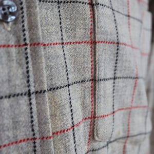 May include: Close-up of a gray plaid shirt with red and black grid lines. The shirt has a small pocket and dark buttons. The fabric appears to be a textured wool or wool blend.