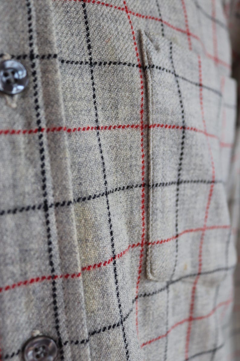 May include: Close-up of a gray plaid shirt with red and black grid lines. The shirt has a small pocket and dark buttons. The fabric appears to be a textured wool or wool blend.