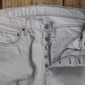 May include: A pair of light gray denim jeans with a button fly and five pockets. The jeans are unwashed and have a raw, unfinished look.