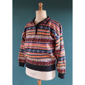 May include: A patterned sweater with a collared neckline and button closure. The sweater has a black collar, cuffs, and hem, with a vibrant mix of orange, red, blue, and purple geometric patterns. The sweater is displayed on a mannequin.