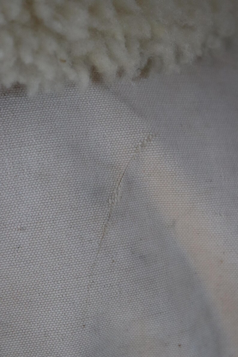 May include: Close-up of a textured, off-white fabric with a visible seam and a fluffy, cream-colored material at the top. The fabric has a subtle, speckled appearance.