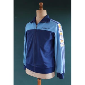 May include: A vintage Adidas track jacket in shades of blue. The jacket features a full zipper, a stand-up collar, and the Adidas logo on the chest. The sleeves have three stripes and a series of colorful patches. The jacket is displayed on a mannequin.