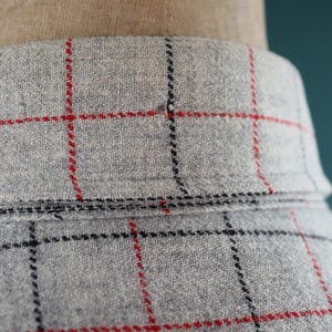 May include: Close-up of a gray fabric with a red and black plaid pattern. The fabric is draped over a mannequin. The collar is visible, and the fabric has a textured appearance.