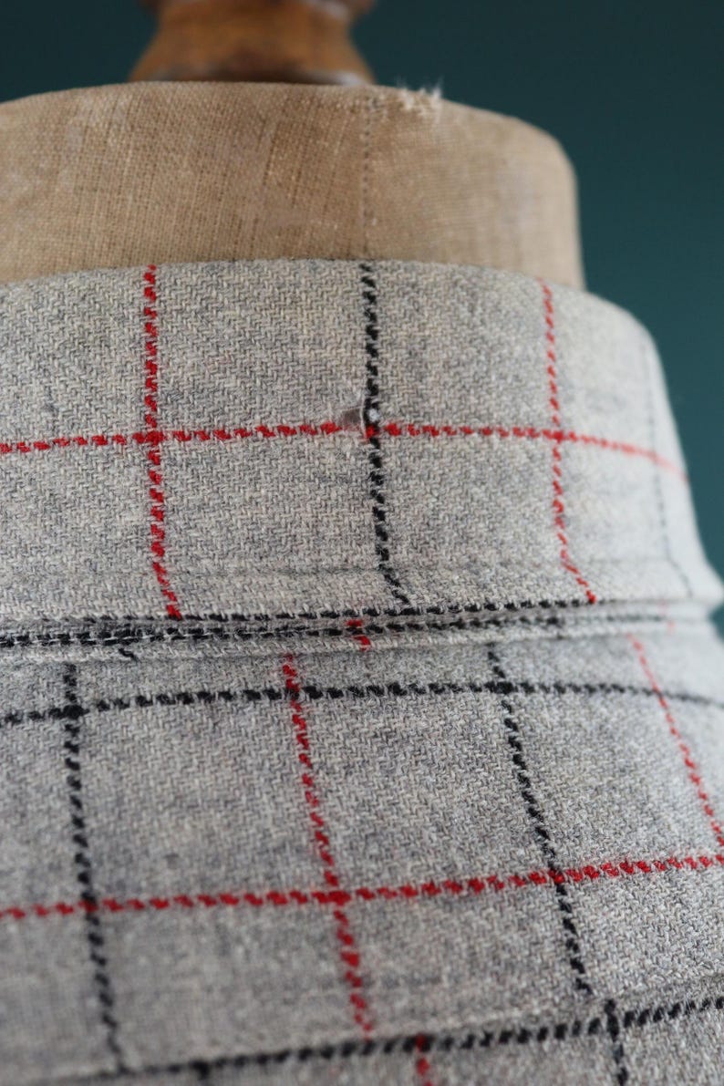 May include: Close-up of a gray fabric with a red and black plaid pattern. The fabric is draped over a mannequin. The collar is visible, and the fabric has a textured appearance.