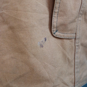 May include: Close-up of a tan fabric with a pocket detail and blue stitching. The fabric has a slightly textured appearance and some dark stains. The image shows a portion of a garment, possibly a jacket or apron.