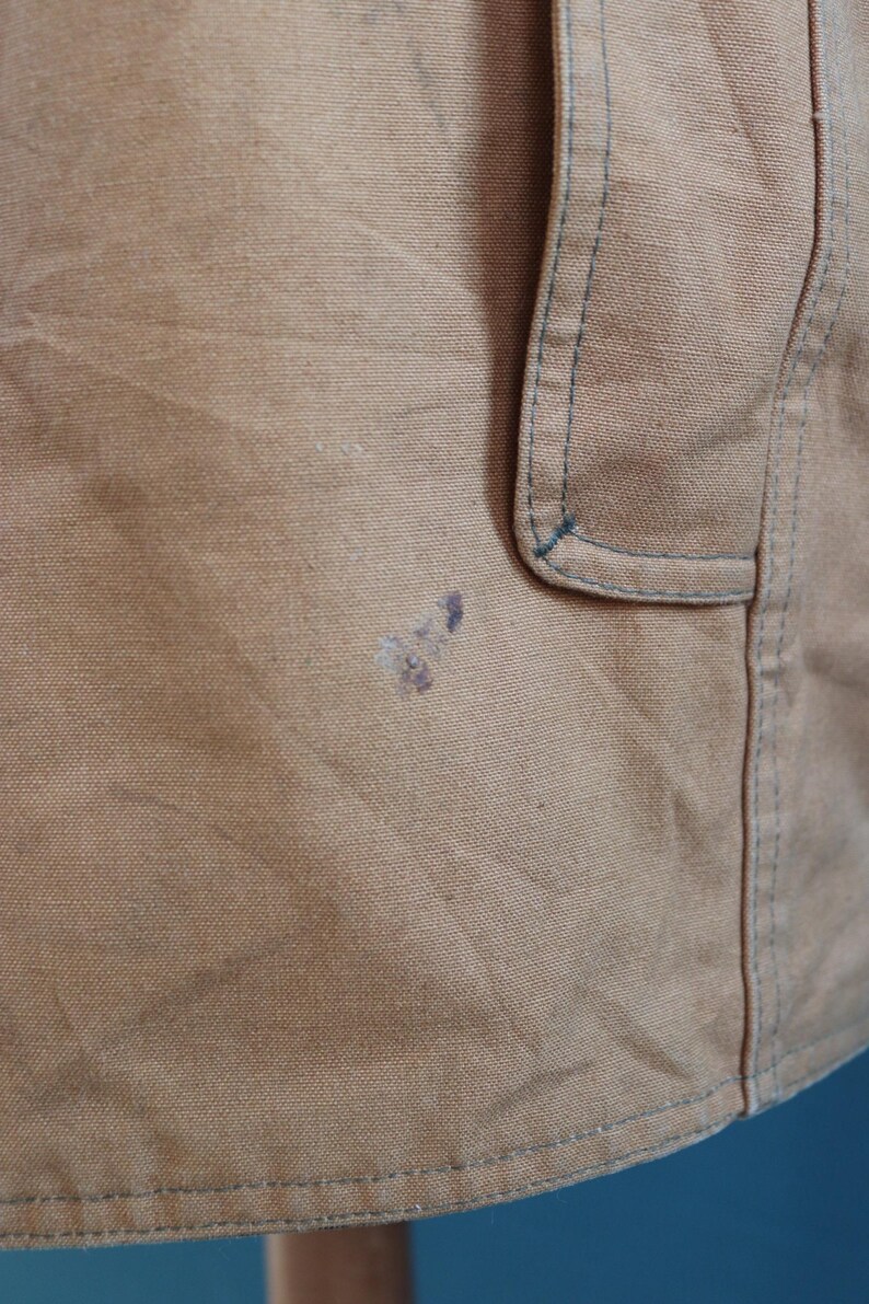 May include: Close-up of a tan fabric with a pocket detail and blue stitching. The fabric has a slightly textured appearance and some dark stains. The image shows a portion of a garment, possibly a jacket or apron.