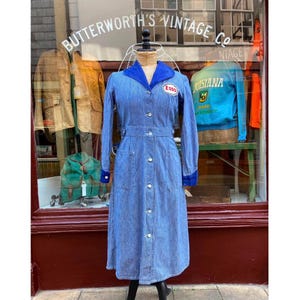 May include: A long-sleeved, button-down dress made of blue and white pinstriped fabric. The dress features a solid blue collar, a white and red Esso logo patch on the chest, two front pockets, and a buttoned waistline, displayed on a mannequin.