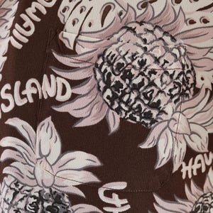 Peut inclure: Chemise boutonn&eacute;e marron avec un imprim&eacute; ananas rose et noir. La chemise a une poche sur le c&ocirc;t&eacute; gauche et le texte "HAWAII ISLAND" imprim&eacute; sur le devant.