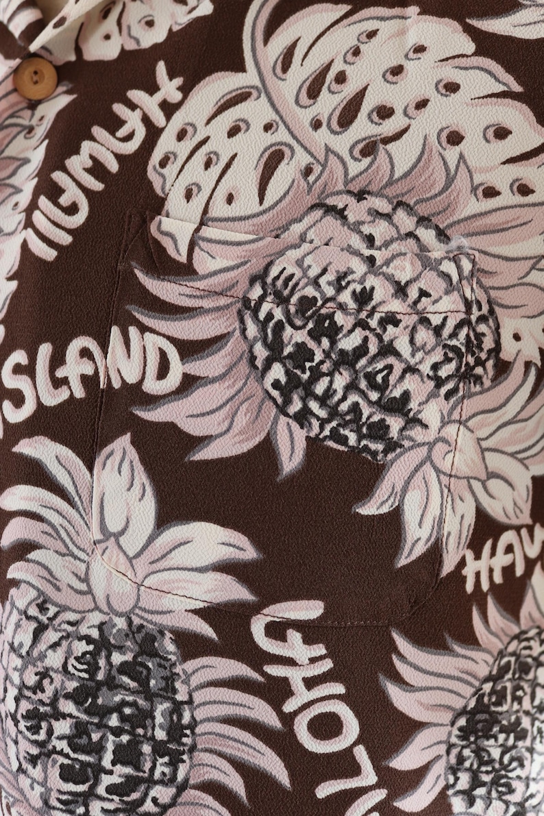 Peut inclure: Chemise boutonn&eacute;e marron avec un imprim&eacute; ananas rose et noir. La chemise a une poche sur le c&ocirc;t&eacute; gauche et le texte "HAWAII ISLAND" imprim&eacute; sur le devant.