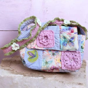 May include: A patchwork handbag with a blue and white gingham pattern, featuring floral and butterfly fabric panels. The bag has crochet details in pink and green, with a matching strap and flower accents. The bag is approximately 10 inches wide.