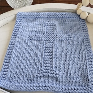 May include: A light blue knitted square with a cross design. The square has a textured appearance with a detailed border. The cross is centered and stands out against the background. The item is likely a decorative piece or a small blanket.