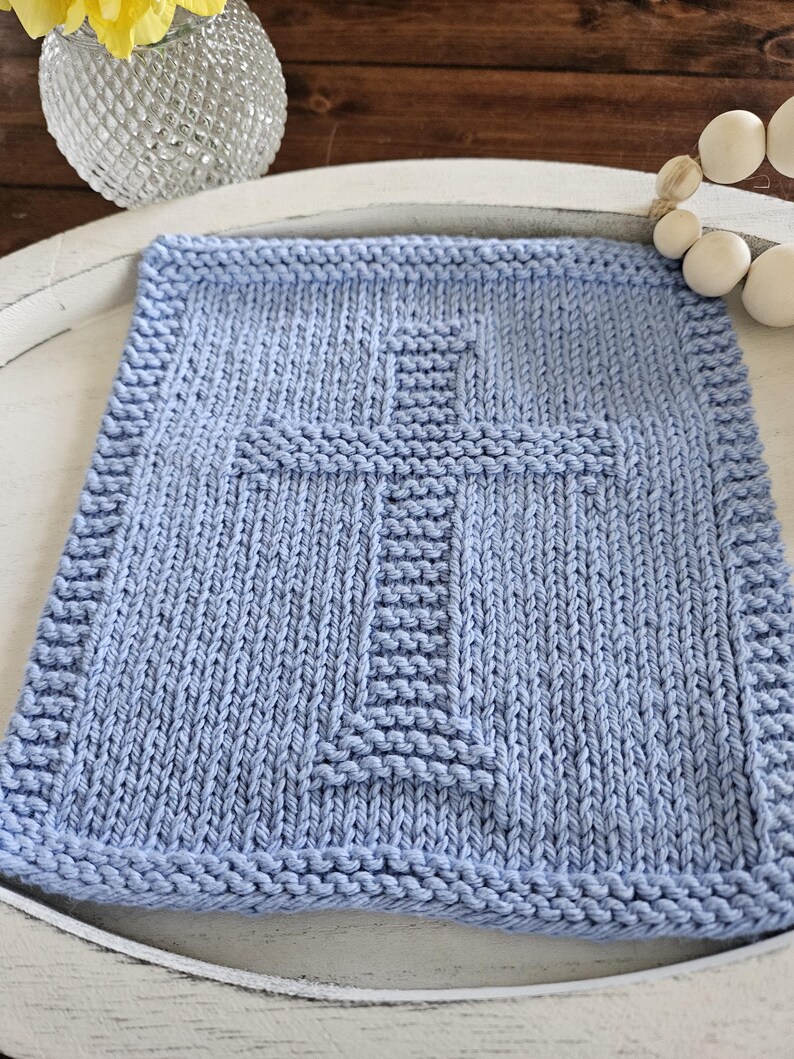 May include: A light blue knitted square with a cross design. The square has a textured appearance with a detailed border. The cross is centered and stands out against the background. The item is likely a decorative piece or a small blanket.