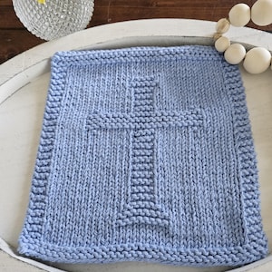 May include: A light blue, square, knitted washcloth with a cross design in the center. The washcloth is placed on a white, oval tray. A vase with yellow flowers and a string of wooden beads are in the background.