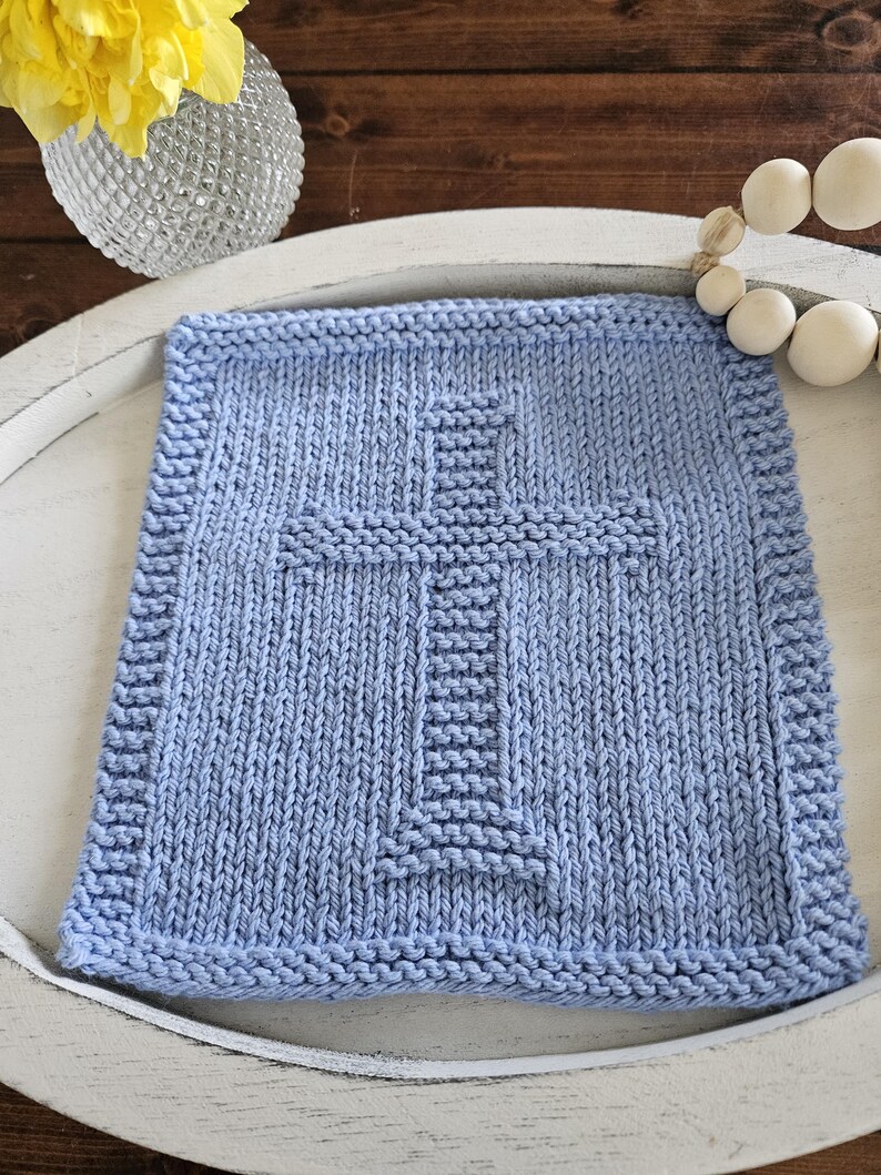 May include: A light blue, square, knitted washcloth with a cross design in the center. The washcloth is placed on a white, oval tray. A vase with yellow flowers and a string of wooden beads are in the background.
