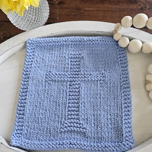 May include: A light blue knitted square with a cross design. The cross is centered and made with a different stitch pattern. The square is on a white tray with a wooden background. A vase with yellow flowers and a string of white beads are also in the image.