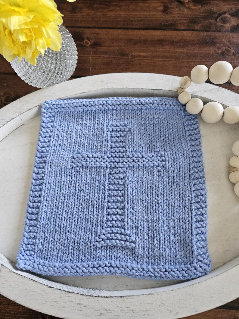 May include: A light blue knitted square with a cross design. The cross is centered and made with a different stitch pattern. The square is on a white tray with a wooden background. A vase with yellow flowers and a string of white beads are also in the image.