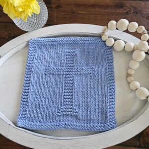 May include: A square, light blue knitted washcloth with a cross design in the center. The washcloth is on a white, oval-shaped tray. A string of white wooden beads and a vase of yellow flowers are also in the image.