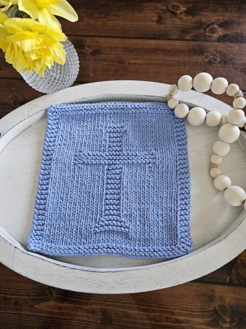 May include: A square, light blue knitted washcloth with a cross design in the center. The washcloth is on a white, oval-shaped tray. A string of white wooden beads and a vase of yellow flowers are also in the image.