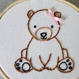 May include: Embroidered brown bear with a pink bow sitting on a white fabric background. The bear has black eyes and a black nose.