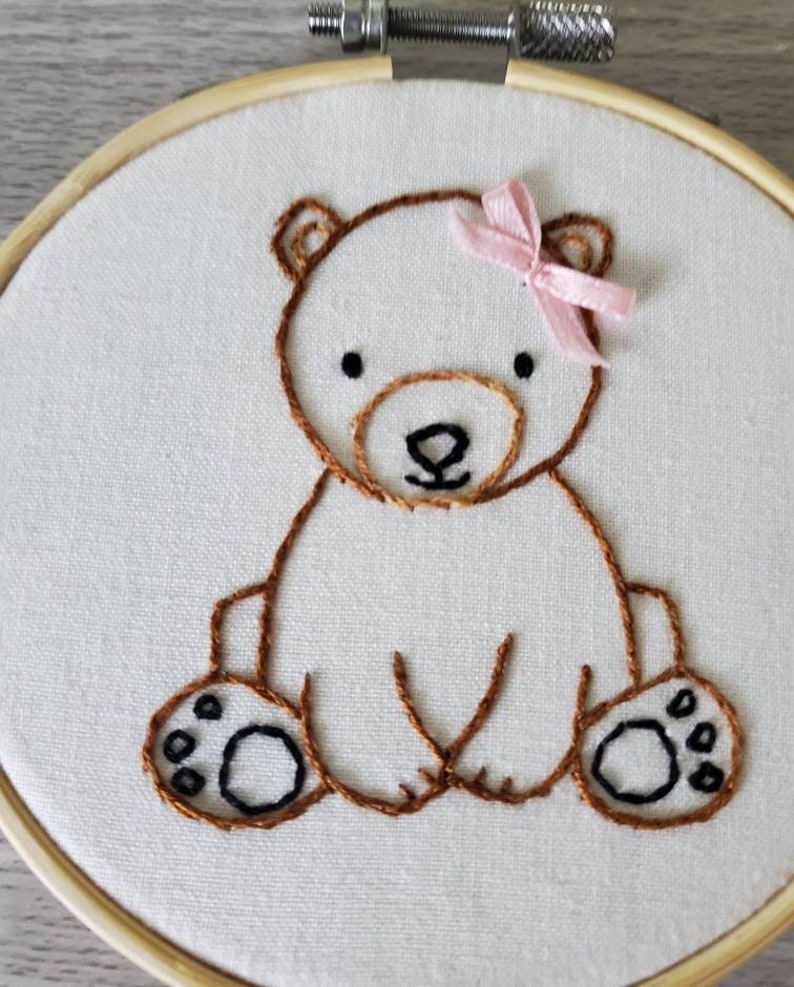 May include: Embroidered brown bear with a pink bow sitting on a white fabric background. The bear has black eyes and a black nose.