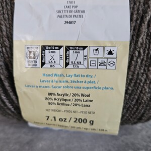 K&ouml;nnte beinhalten: Ein Kn&auml;uel dunkelgraues Garn mit einem Produktetikett, das den Text CAKE POP, 80% Acryl / 20% Wolle und ein Nettogewicht von 200 g enth&auml;lt. Das Etikett zeigt Pflegehinweise f&uuml;r Handw&auml;sche und flaches Trocknen sowie Symbole f&uuml;r Strick- und H&auml;kelmaschen.
