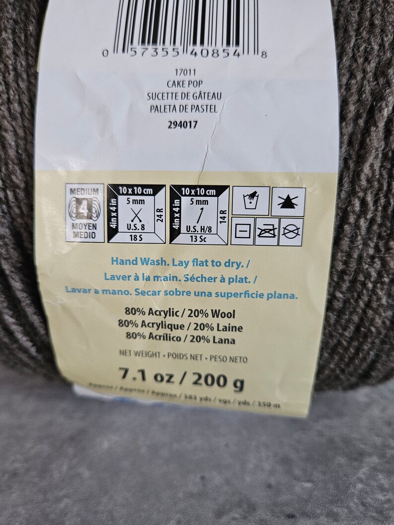 K&ouml;nnte beinhalten: Ein Kn&auml;uel dunkelgraues Garn mit einem Produktetikett, das den Text CAKE POP, 80% Acryl / 20% Wolle und ein Nettogewicht von 200 g enth&auml;lt. Das Etikett zeigt Pflegehinweise f&uuml;r Handw&auml;sche und flaches Trocknen sowie Symbole f&uuml;r Strick- und H&auml;kelmaschen.