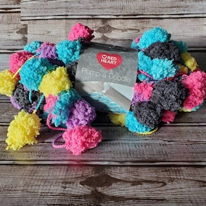 May include: A skein of Red Heart Pom-a-Doodle yarn in a multi-colored pom-pom style. The yarn is in shades of pink, yellow, blue, purple, and gray.