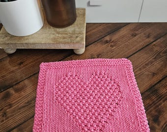 May include: A pink, square, knitted washcloth with a heart design in the center. The washcloth is made of a soft, textured yarn and has a ribbed border. The heart is created with a raised stitch pattern.