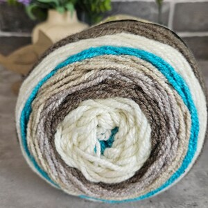 K&ouml;nnte beinhalten: Ein rundes Garnkn&auml;uel mit konzentrischen Farbringen in den T&ouml;nen Creme, Hellbeige, Mittelbraun, Dunkelbraun und einem leuchtenden T&uuml;rkisblau. Das Garn hat eine weiche, gedrehte Textur und ist zu einem dichten, kreisf&ouml;rmigen Ball gewickelt.