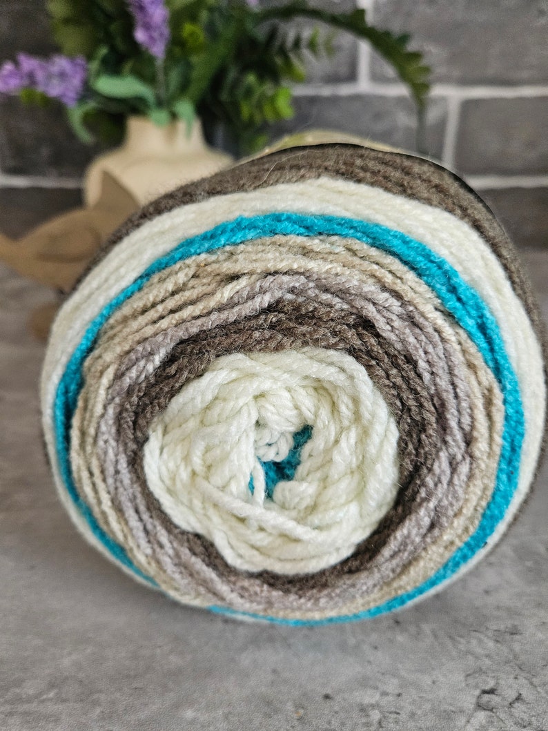 K&ouml;nnte beinhalten: Ein rundes Garnkn&auml;uel mit konzentrischen Farbringen in den T&ouml;nen Creme, Hellbeige, Mittelbraun, Dunkelbraun und einem leuchtenden T&uuml;rkisblau. Das Garn hat eine weiche, gedrehte Textur und ist zu einem dichten, kreisf&ouml;rmigen Ball gewickelt.