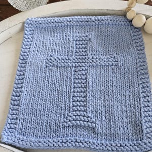 May include: A light blue, square, knitted blanket with a cross design in the center. The blanket has a textured appearance and a thick border. The cross is created with a different stitch pattern, adding dimension to the design. The blanket is likely handmade.