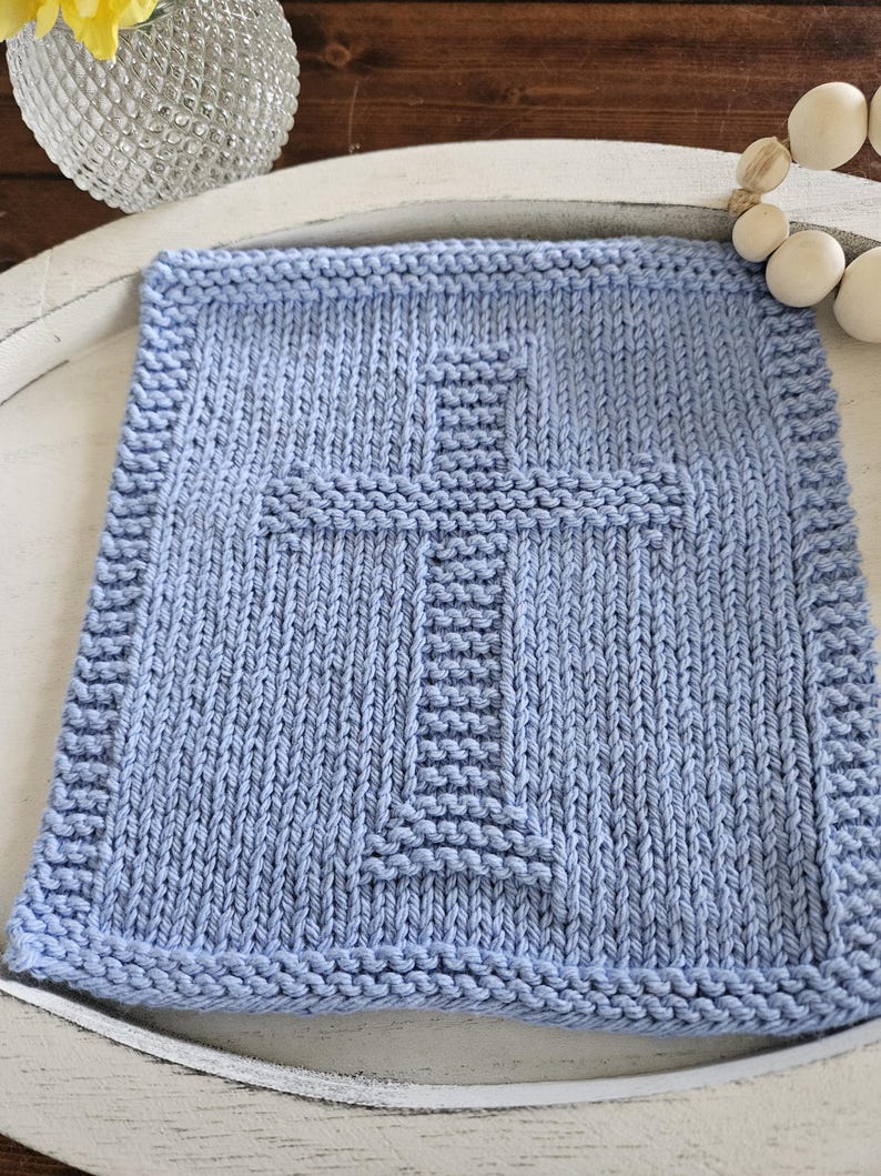 May include: A light blue, square, knitted blanket with a cross design in the center. The blanket has a textured appearance and a thick border. The cross is created with a different stitch pattern, adding dimension to the design. The blanket is likely handmade.