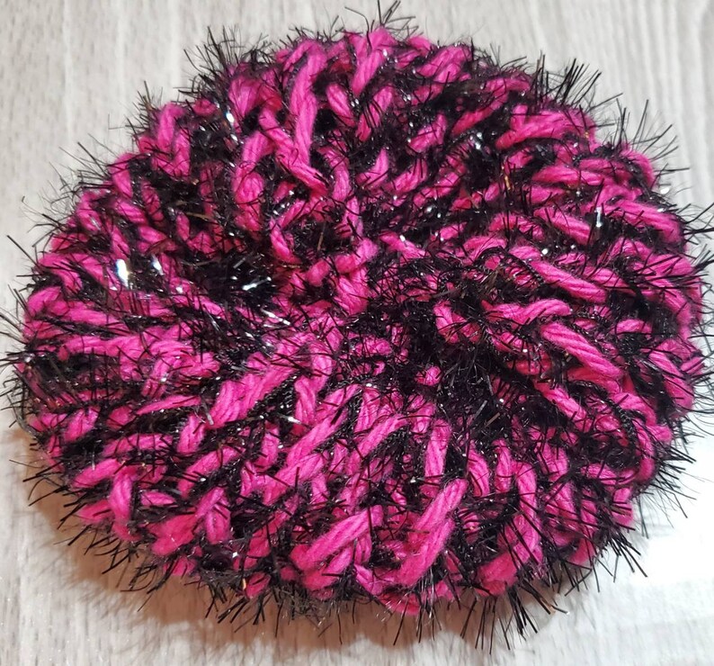 Puede incluir: Una esponja de fregar de hilo rosa y negro con un acabado brillante. La esponja est&aacute; hecha de hilo esponjoso y es perfecta para limpiar platos u otras superficies.