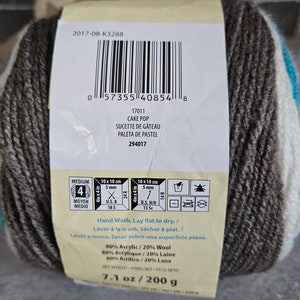 K&ouml;nnte beinhalten: Ein Garnkn&auml;uel mit der Aufschrift Cake Pop, bestehend aus 80 % Acryl und 20 % Wolle. Das Etikett zeigt einen Barcode, Pflegehinweise f&uuml;r Handw&auml;sche und ein Nettogewicht von 200 g. Das Garn hat die St&auml;rke 4 und enth&auml;lt Angaben zur Maschenprobe.