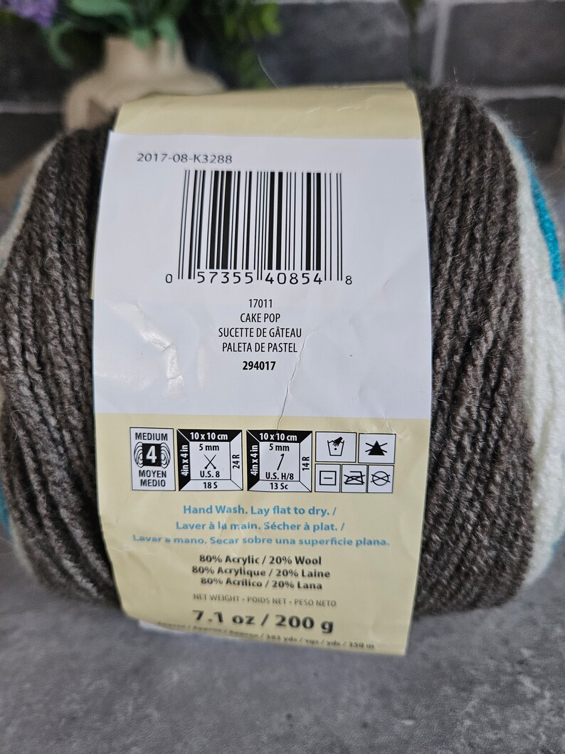 K&ouml;nnte beinhalten: Ein Garnkn&auml;uel mit der Aufschrift Cake Pop, bestehend aus 80 % Acryl und 20 % Wolle. Das Etikett zeigt einen Barcode, Pflegehinweise f&uuml;r Handw&auml;sche und ein Nettogewicht von 200 g. Das Garn hat die St&auml;rke 4 und enth&auml;lt Angaben zur Maschenprobe.