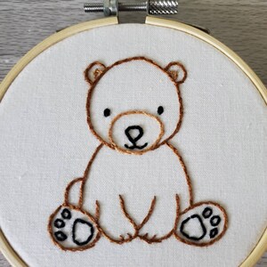 May include: Embroidered brown bear sitting on a white fabric background. The bear has black eyes and a brown nose. The bear is surrounded by a wooden hoop.