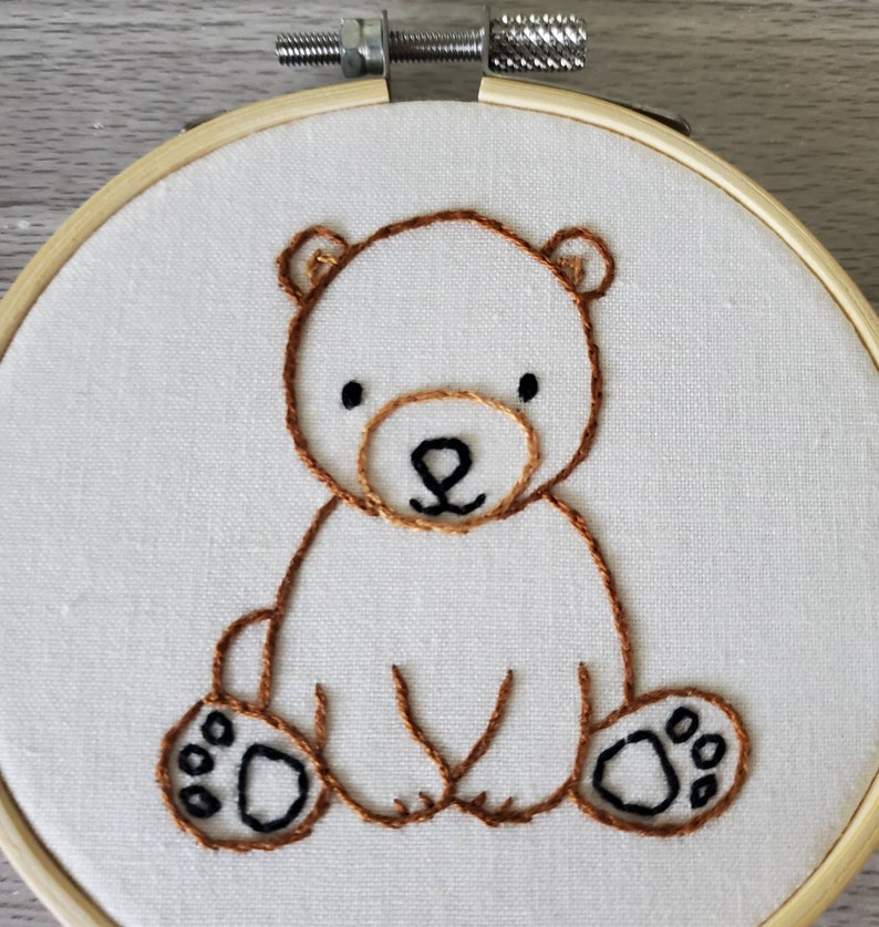 May include: Embroidered brown bear sitting on a white fabric background. The bear has black eyes and a brown nose. The bear is surrounded by a wooden hoop.