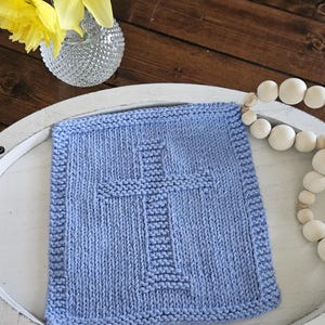 May include: A light blue knitted square with a cross design in the center. The square is placed on a white tray, with a vase of yellow flowers and a string of white beads in the background.