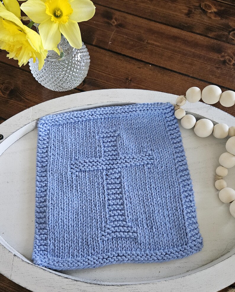 May include: A light blue knitted square with a cross design in the center. The square is placed on a white tray, with a vase of yellow flowers and a string of white beads in the background.