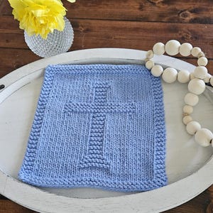 May include: A light blue, square, knitted washcloth with a cross design. The washcloth is placed on a white tray with a wooden background. A vase with yellow flowers and a beaded garland are also in the image.