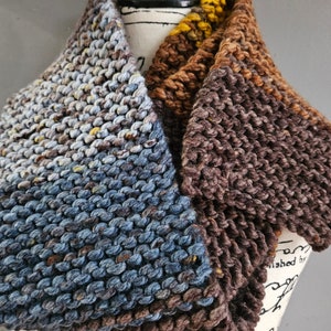 Puede incluir: Una bufanda de cuello de punto en tonos marrones, azules y amarillos. La bufanda está hecha con un hilo grueso y tiene un patrón de punto texturizado.