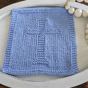 May include: A light blue, square, knitted blanket with a cross design in the center. The blanket has a textured appearance and a decorative border. The blanket is placed on a white tray, with a string of white beads in the background.