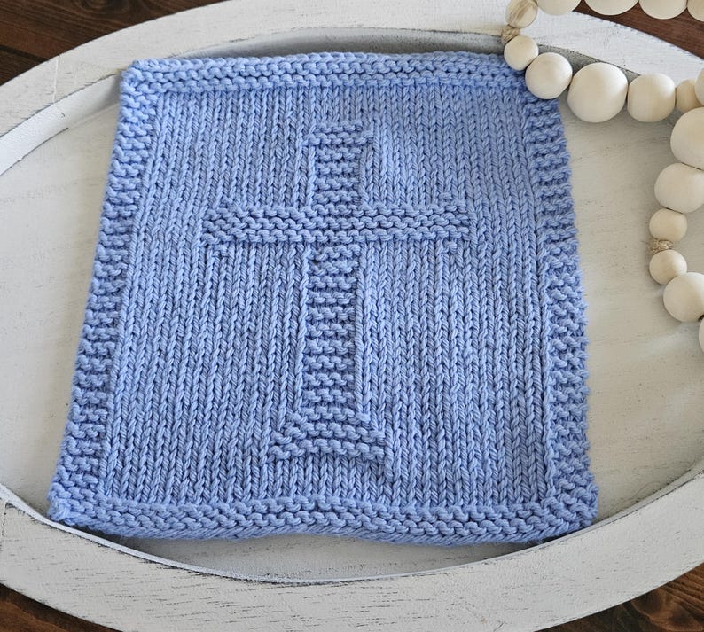 May include: A light blue, square, knitted blanket with a cross design in the center. The blanket has a textured appearance and a decorative border. The blanket is placed on a white tray, with a string of white beads in the background.