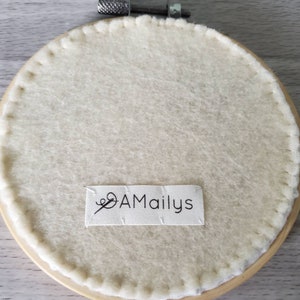 May include: The back of a round embroidery hoop with a white felt backing. The hoop is made of wood and has a metal screw closure. A white label with the text "AMailys" is attached to the felt.