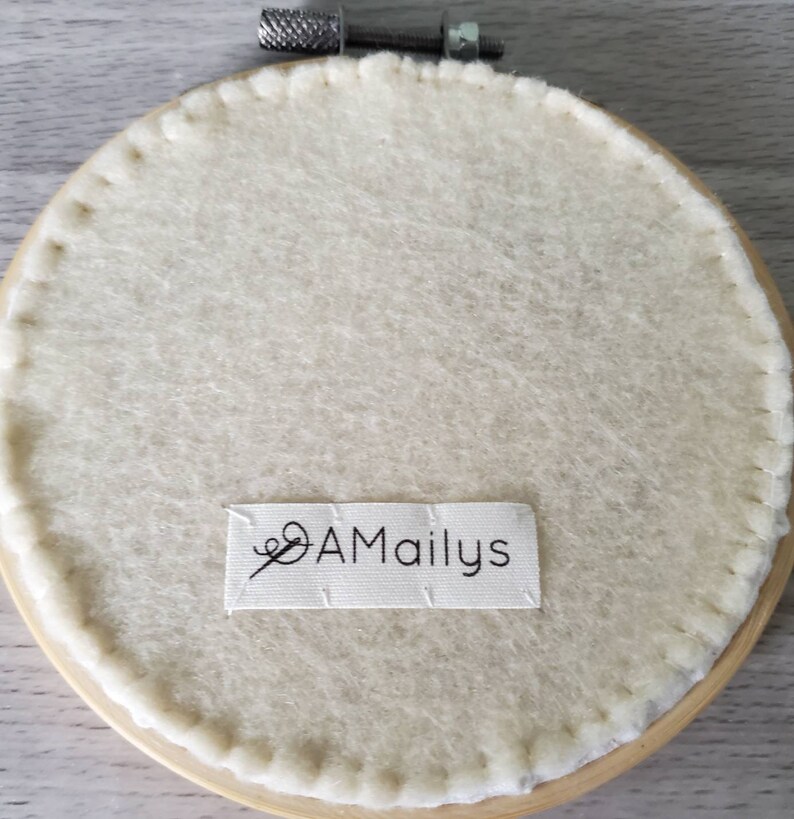 May include: The back of a round embroidery hoop with a white felt backing. The hoop is made of wood and has a metal screw closure. A white label with the text "AMailys" is attached to the felt.