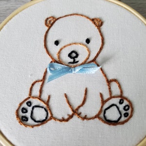May include: Embroidered brown bear with a blue bow tie on a white fabric background. The bear is sitting with its paws in front of it.
