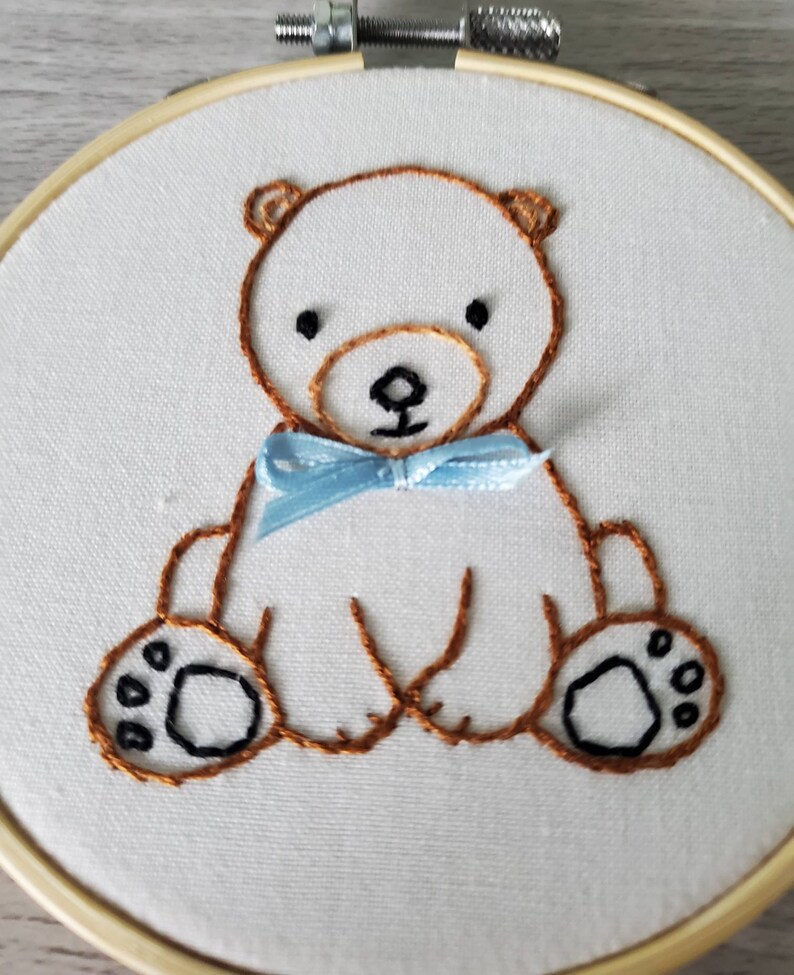 May include: Embroidered brown bear with a blue bow tie on a white fabric background. The bear is sitting with its paws in front of it.
