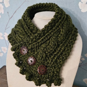 Puede incluir: Un cuello de punto de ganchillo verde oscuro con un patrón de trenza y tres botones de madera. El cuello tiene un borde festoneado.