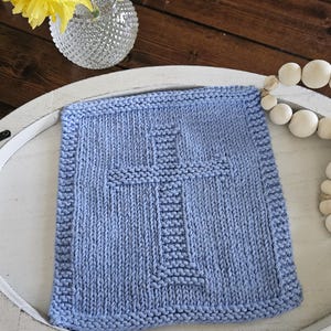 May include: A square, light blue knitted washcloth with a prominent cross design in the center. The washcloth has a textured appearance and a ribbed border. The item is displayed on a white tray, with a vase of yellow flowers in the background.