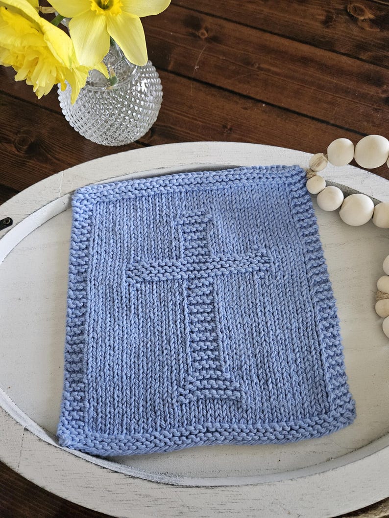 May include: A square, light blue knitted washcloth with a prominent cross design in the center. The washcloth has a textured appearance and a ribbed border. The item is displayed on a white tray, with a vase of yellow flowers in the background.